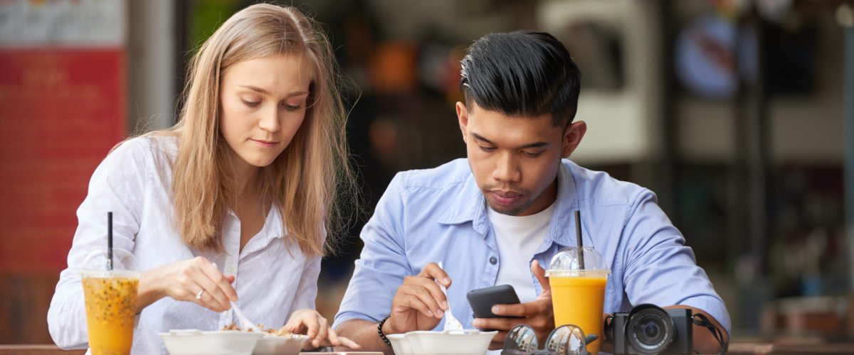 Young couple of tourists eating Asian street food, man using his smartphone