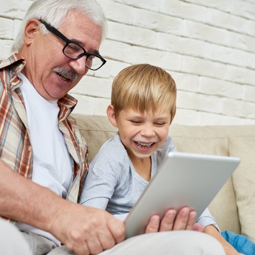 Portrait of happy senior man using digital tablet watching videos with grandson both laughing sitting on couch at home